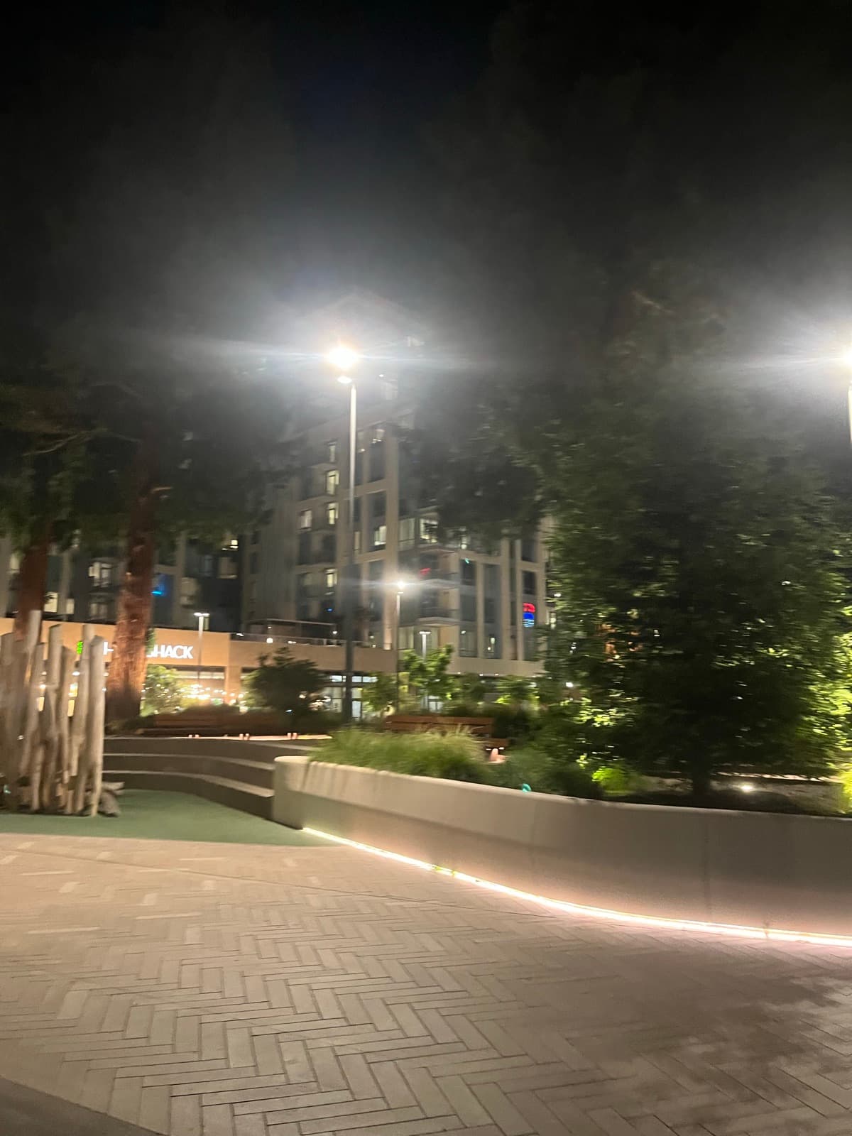 A quiet plaza at night, planters and benches lit under a tall apartment building