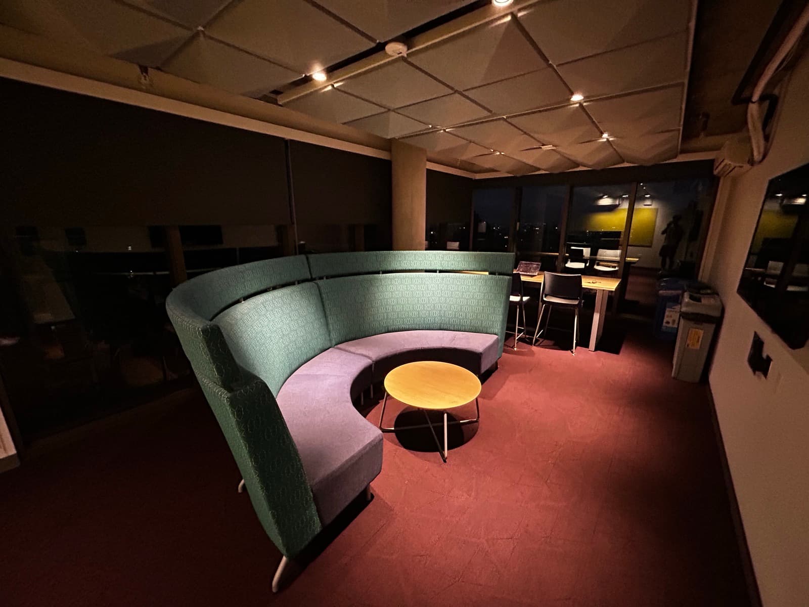 An empty study lounge late at night: a curved green booth around a small round table, the city dark through the windows behind