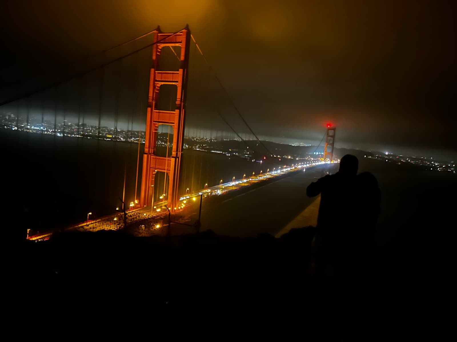 The Golden Gate Bridge lit up at night, the city glowing behind it, a friend silhouetted against the lights