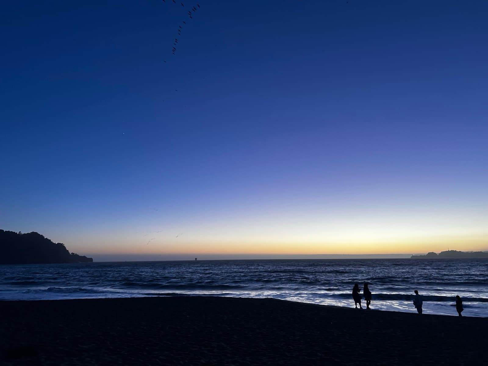 A dark beach at dusk, a few people at the water’s edge, cliffs and a pale orange horizon